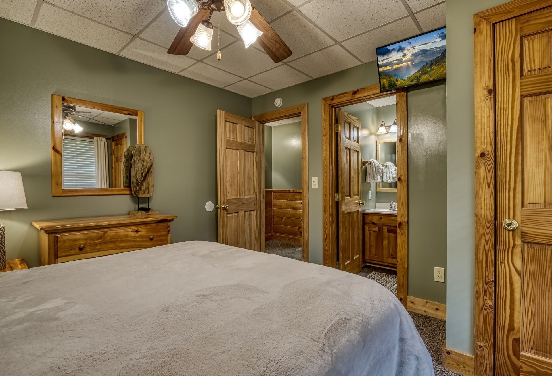 Une chambre à coucher avec un lit, une armoire en bois, un miroir, un ventilateur de plafond et un téléviseur fixé au mur.