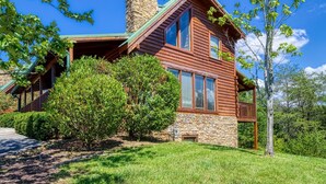 A two-story log cabin with a stone chimney, surrounded by lush greenery and a clear blue sky. - Ern813 - Heavenly View- Great Location! Close To All The Action! 6 Bedroom Cabin by RedAwning (Sevierville)