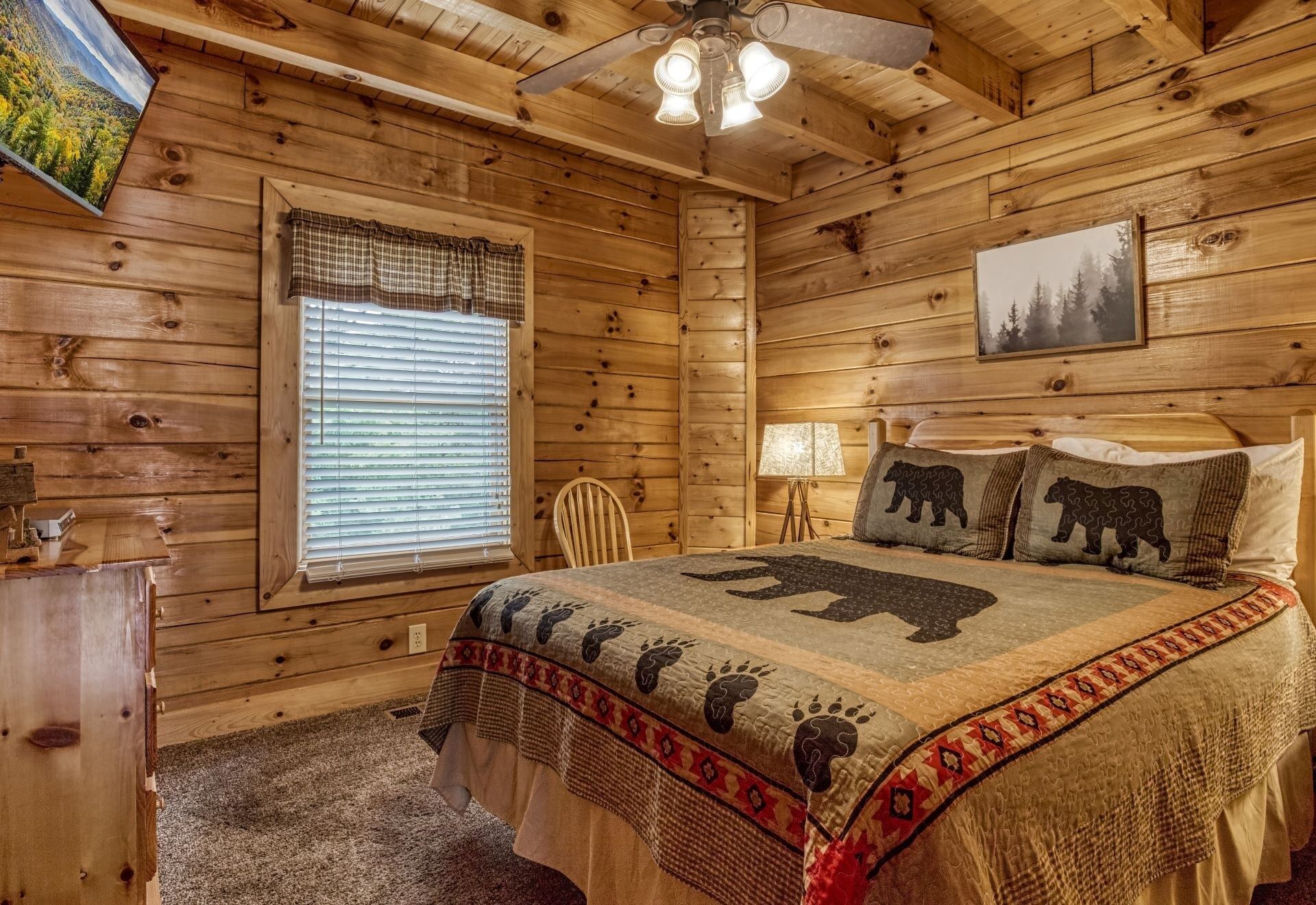 A cozy log cabin bedroom with a bed, a window with blinds, a ceiling fan, and a bear-themed pillow.