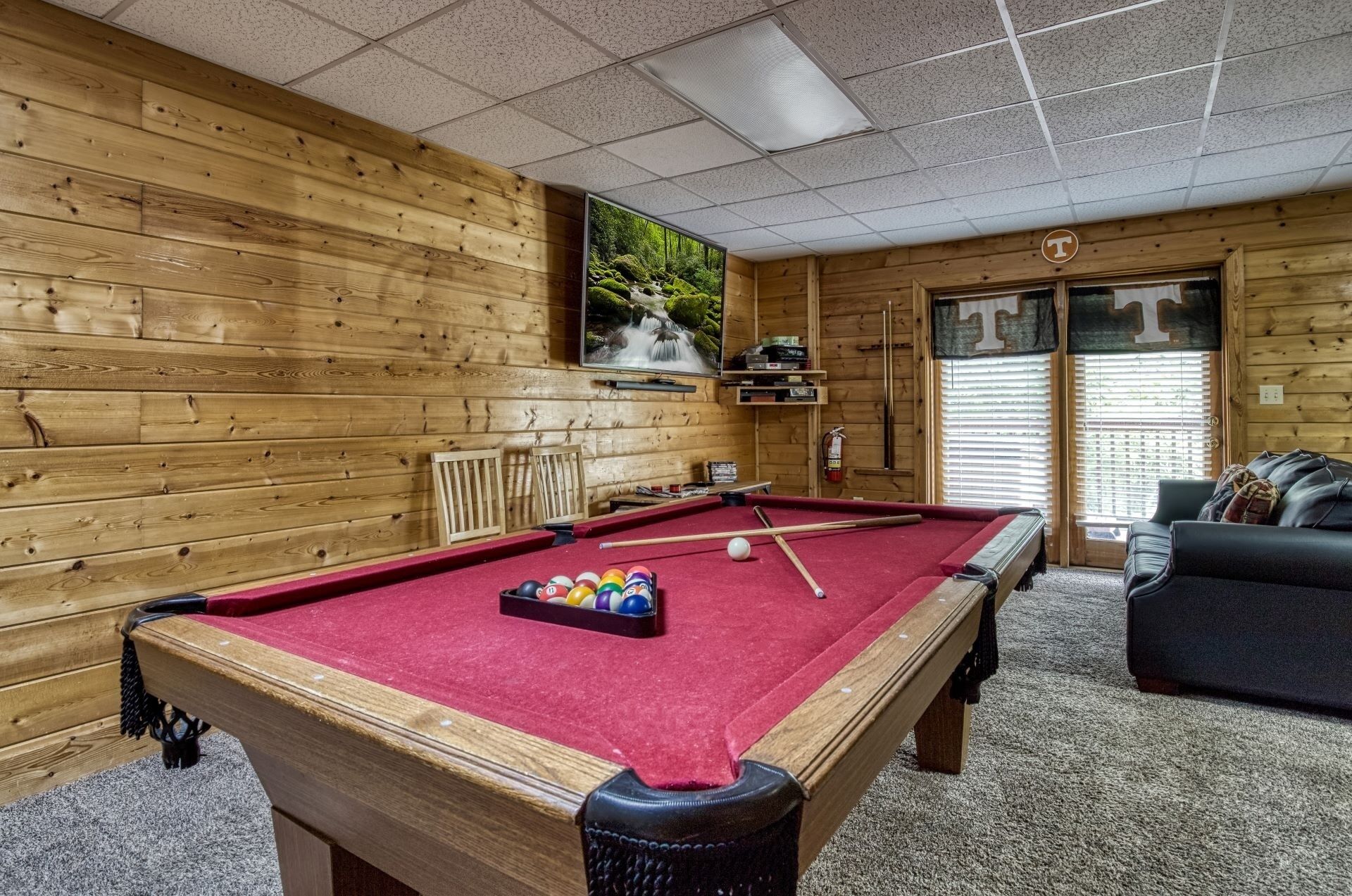 A pool table with balls, a pool cue, and a red felt surface. There is a wooden wall with a window showing a forest scene. A black couch and a door with a 'T' sign are visible.