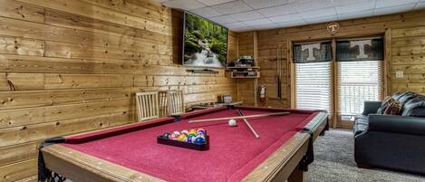 A pool table with balls, a pool cue, and a red felt surface. There is a wooden wall with a window showing a forest scene. A black couch and a door with a 'T' sign are visible.