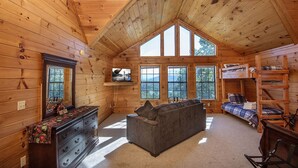 A cozy cabin interior with a loft bed, a sofa, a TV mounted on the wall, and large windows offering a view of the outdoors. - Dunder Mountain Views Cabin Retreat 3 Bedroom Cabin by RedAwning (Sevierville)