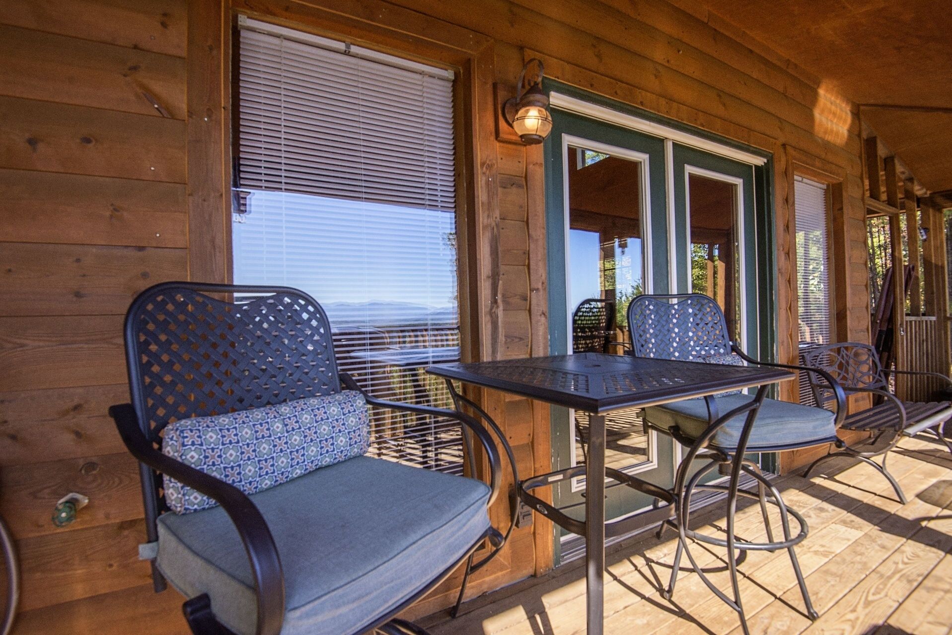 Une cabane en bois dotée d’un porche couvert, avec deux tables et des chaises en métal, une vue sur les montagnes et une porte verte.