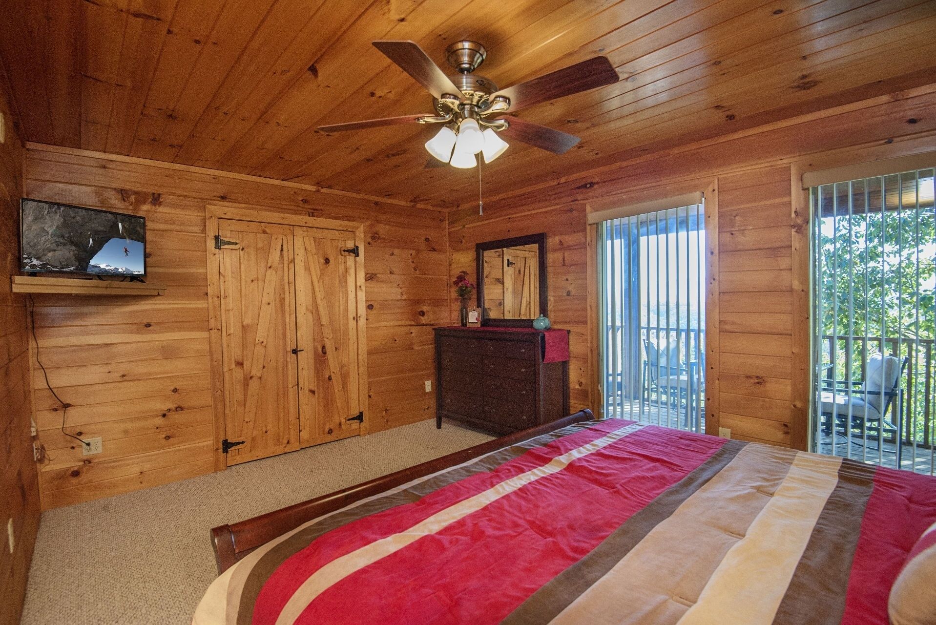 A wooden cabin bedroom with a bed, a ceiling fan, a dresser, a mirror, and a TV on the wall.