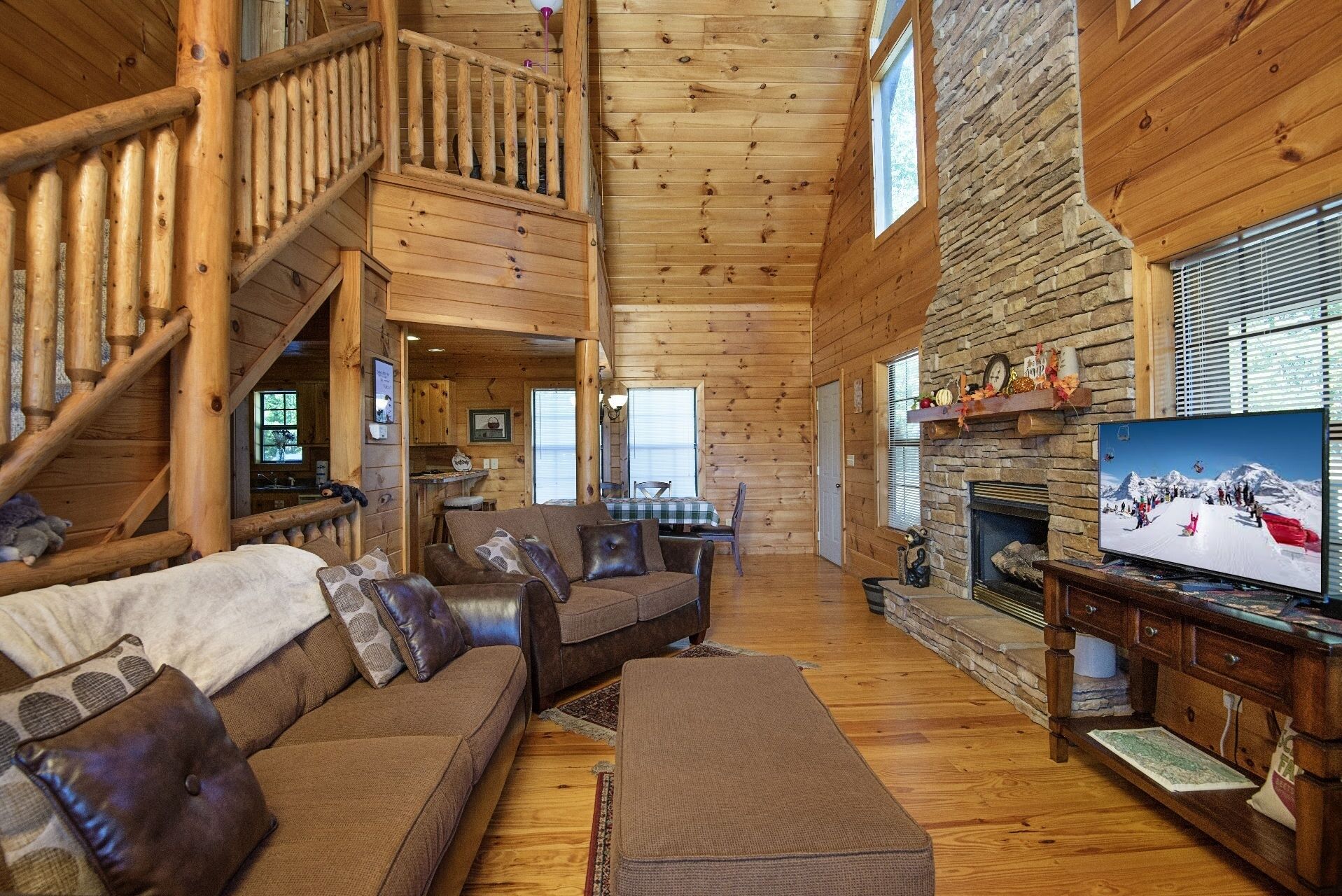 A log cabin interior with a wooden staircase, a stone fireplace, a television, and a cozy seating area.
