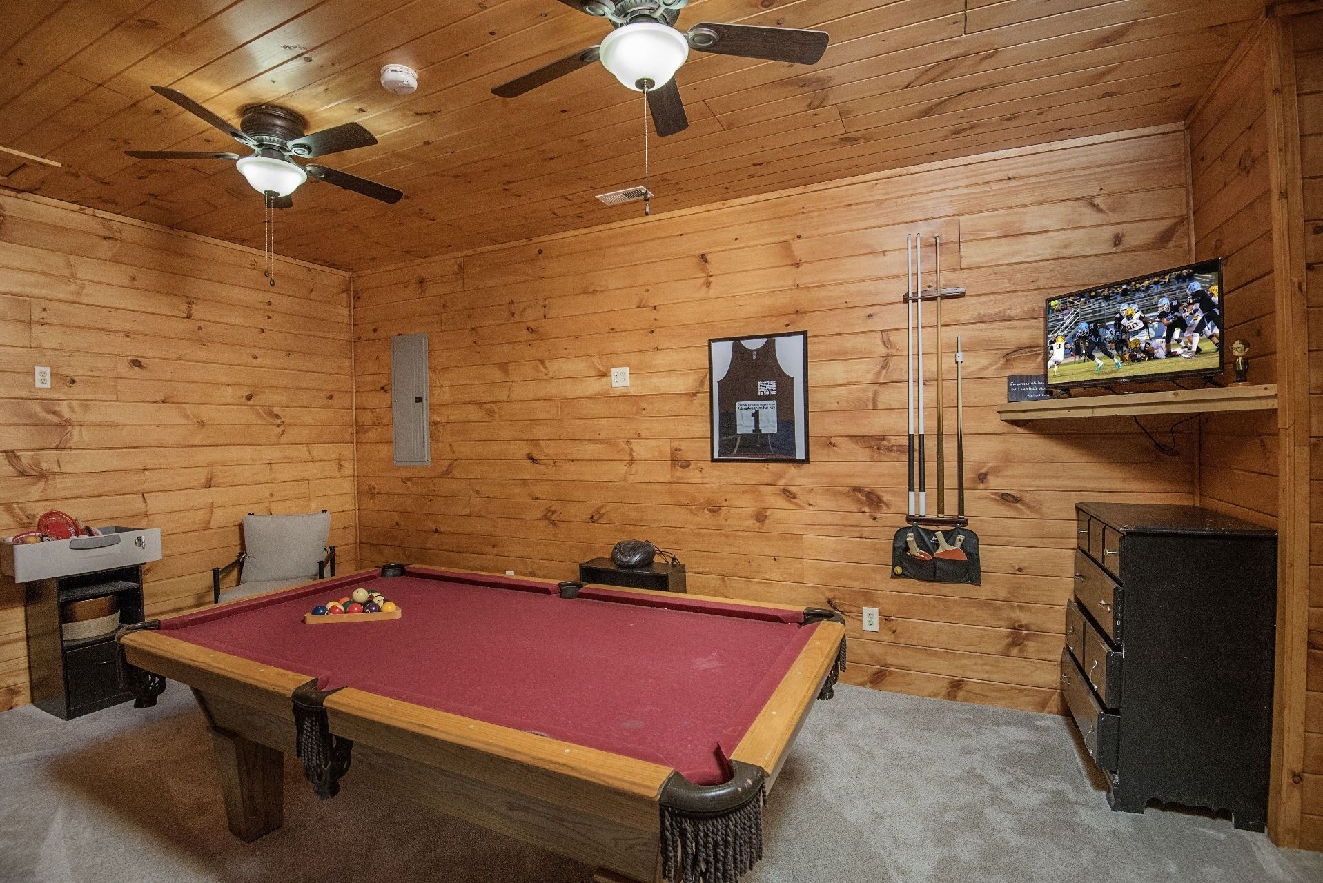 A wooden-paneled room with a pool table, ceiling fans, a television, and a framed jersey on the wall.