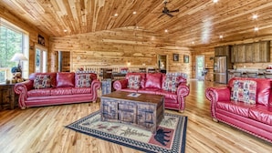 Ein geräumiges Blockhäuschen mit Holzbalkendecken, roten Ledersofas mit gemusterten Kissen, einem zentralen Holztisch und einer Küchenzeile im Hintergrund.