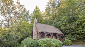 A log cabin with a stone chimney, surrounded by lush greenery and a paved driveway. - Er36 - Cozy Creekside Great Location- Close To Town 1 Bedroom Cabin by RedAwning (Pigeon Forge)