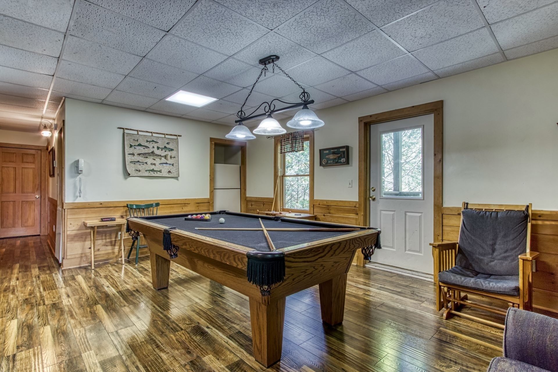 A pool table in a room with wooden flooring, a ceiling with fluorescent lights, a door with a window, and a chair with a cushion.