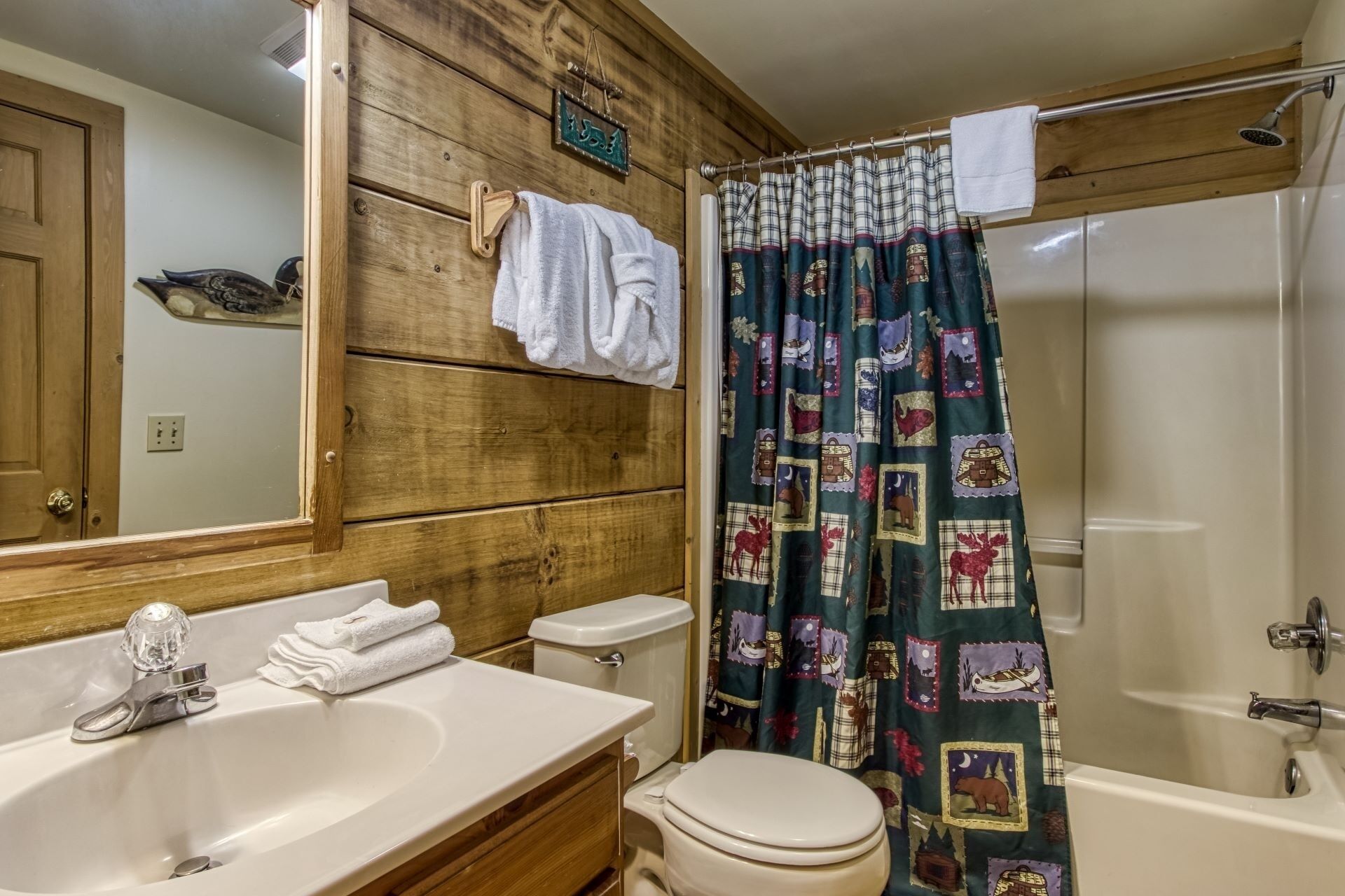 A bathroom with wooden paneling, a sink with a mirror, a toilet, and a shower with a patterned curtain.