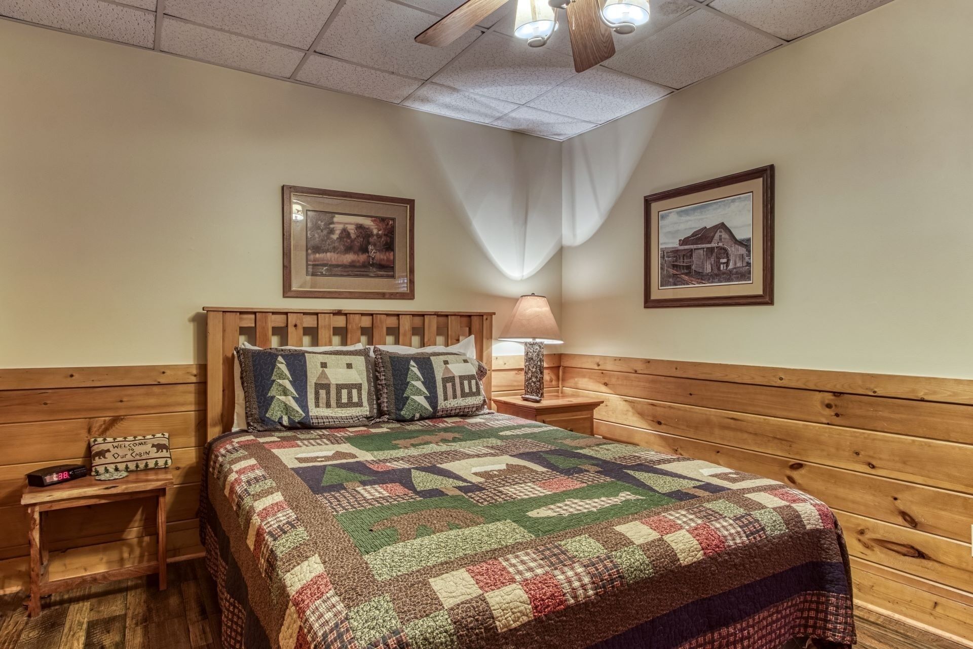 A cozy cabin-style room with a wooden headboard, a quilted bedspread, a nightstand with a lamp, and two framed pictures on the walls.
