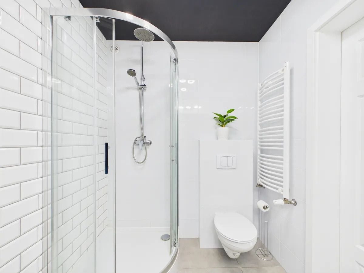Un baño moderno con ducha de vidrio, paredes de azulejos blancos, inodoro blanco y un radiador.