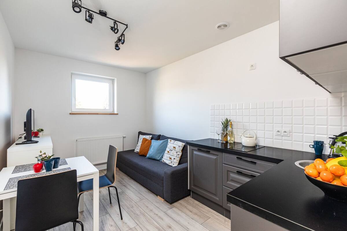 Una cocina moderna con encimera negra, salpicadero blanco de azulejos y sofá gris con cojines estampados.