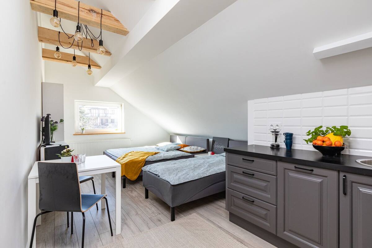 Un dormitorio moderno y funcional con cama, mesita de noche y escritorio con silla. Una ventana ofrece luz natural, y se aprecia una cocina con armarios y encimera.