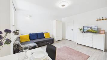 Una sala moderna con un sofá, una mesa de comedor con platos y vasos, un televisor sobre un mueble bajo y una puerta que da a otra habitación.