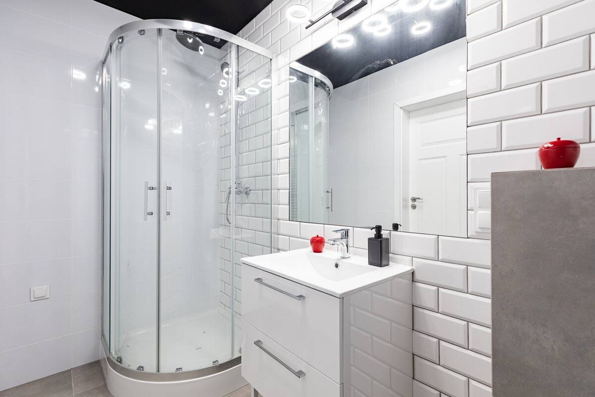 Un baño moderno con ducha de vidrio, un tocador blanco con un objeto rojo encima y un espejo grande sobre el lavabo.