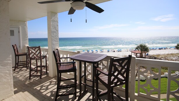 A balcony with a ceiling fan, wooden chairs, and a table. The balcony overlooks a beach with the ocean and beach umbrellas.