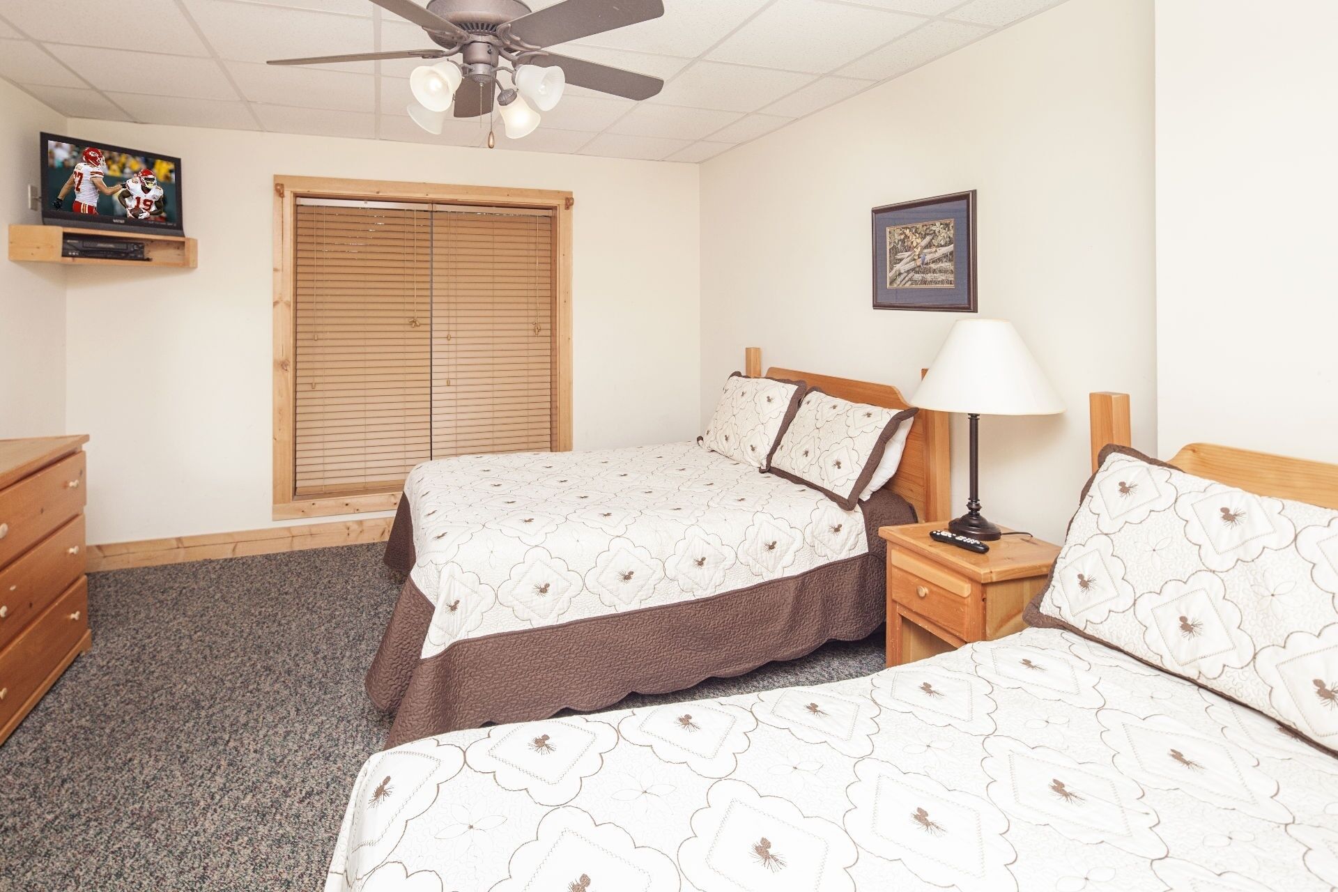 A bedroom with two beds, a ceiling fan, a TV, a window with blinds, a bedside table with a lamp, and a framed picture on the wall.