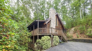 A cabin with a stone chimney, wooden deck, and surrounded by forest. - Er314- Y'all Come Back Now- Great Location- Close To Town 2 Bedroom Cabin by RedAwning (Pigeon Forge)