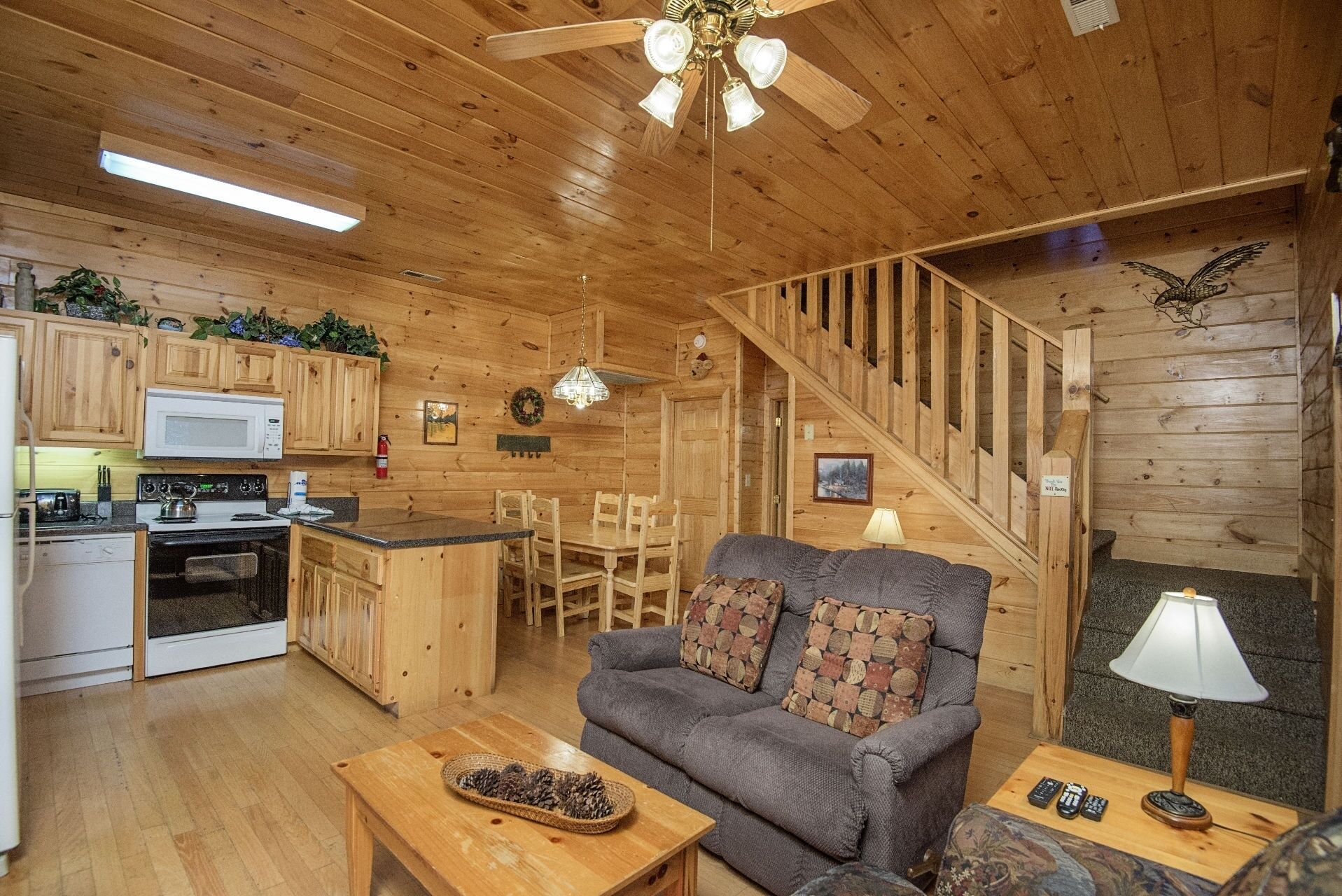 A cozy cabin interior with wooden walls, a kitchen with a microwave, stove, and cabinets, a gray sofa with patterned cushions, a wooden dining table, and a staircase.