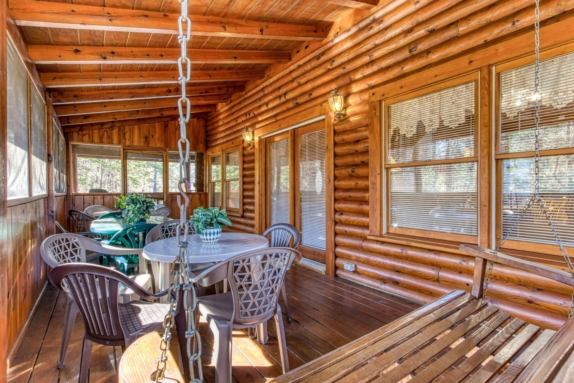 A wooden cabin with a dining area featuring a round table and chairs, large windows with blinds, and a ceiling with exposed beams.