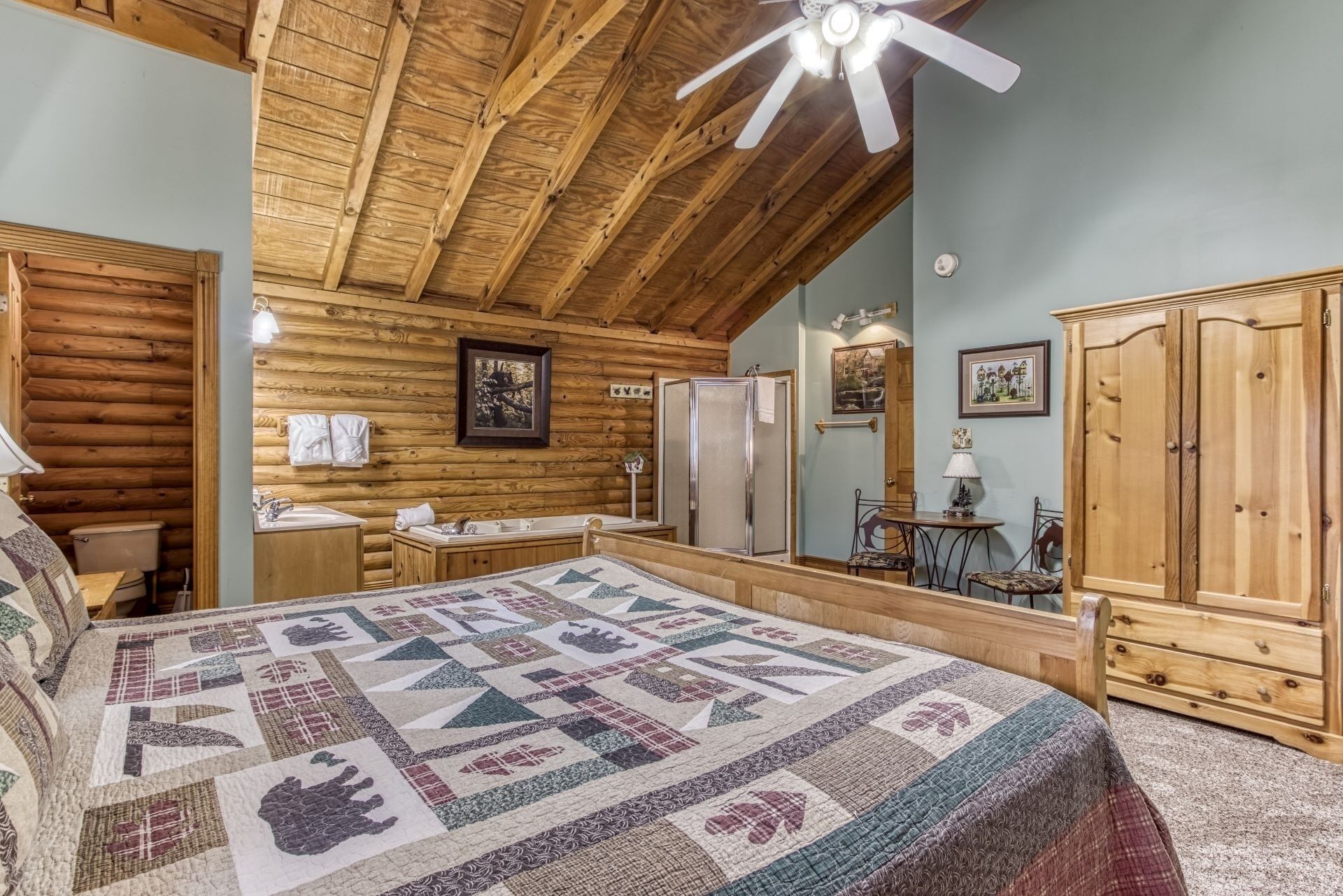 A log cabin bedroom with a large bed, wooden ceiling beams, a ceiling fan, a wooden wardrobe, and a bathroom with a sink and toilet.