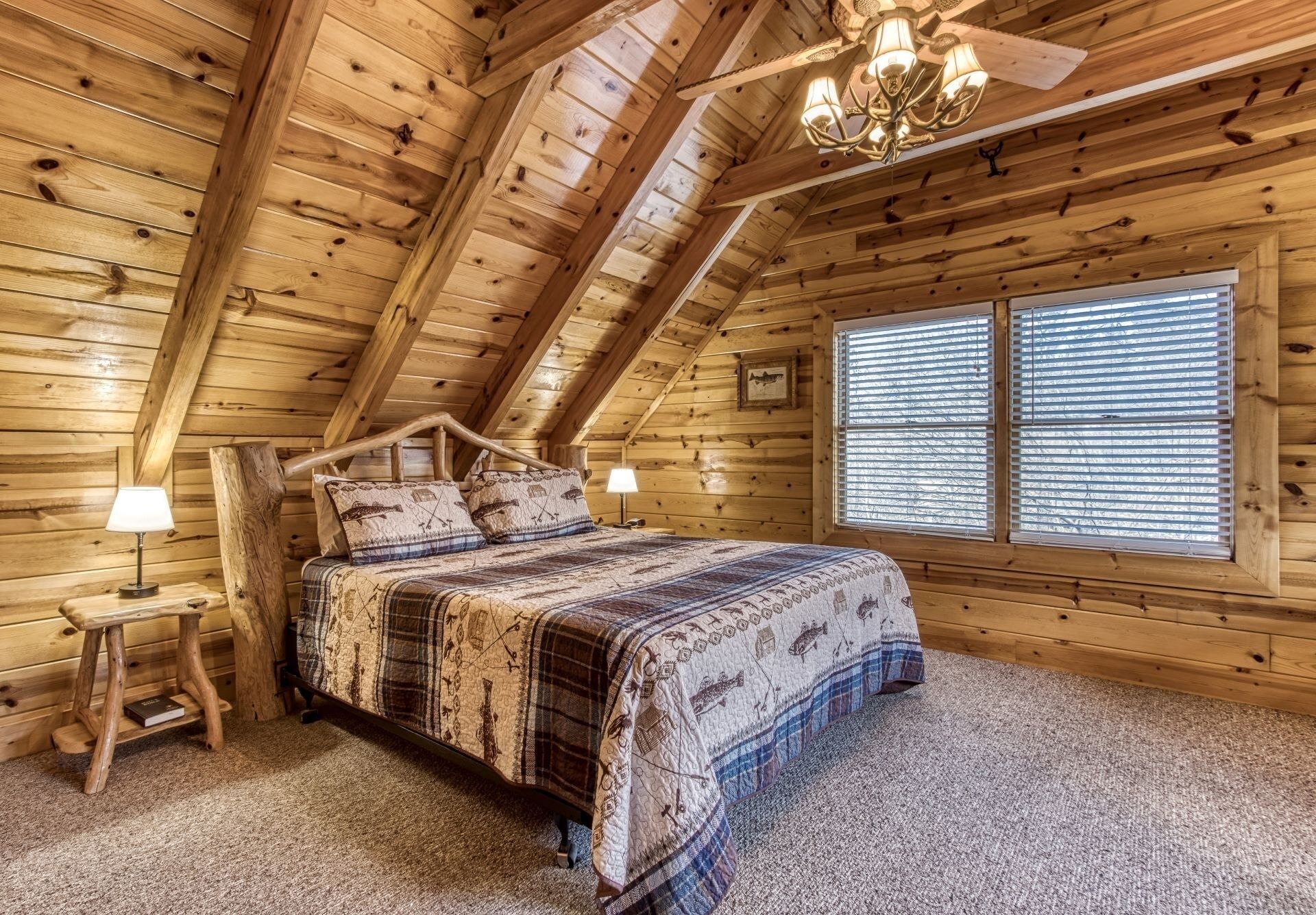 A cozy log cabin bedroom with a bed, two bedside lamps, a chandelier, and a window with blinds.