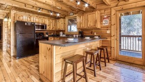 A wooden cabin interior with a kitchen featuring a black refrigerator, a microwave, and a stove. There are wooden stools at the kitchen island, and a window with a view of trees. - The Wildlife Lodge - Great Location! Close To Tanger Outlets! 5 Bedroom Cabin by RedAwning (Sevierville)