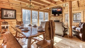 A wooden cabin interior with a dining table set for four, a leather sofa, and a television mounted on a stone wall. - The Wildlife Lodge - Great Location! Close To Tanger Outlets! 5 Bedroom Cabin by RedAwning (Sevierville)