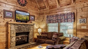 A cozy log cabin interior with a stone fireplace, wooden walls, a comfortable sofa, and a television mounted above the fireplace.