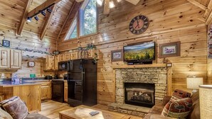 A cozy cabin interior with a stone fireplace, wooden walls, a kitchen with a refrigerator, and a television mounted above the fireplace.