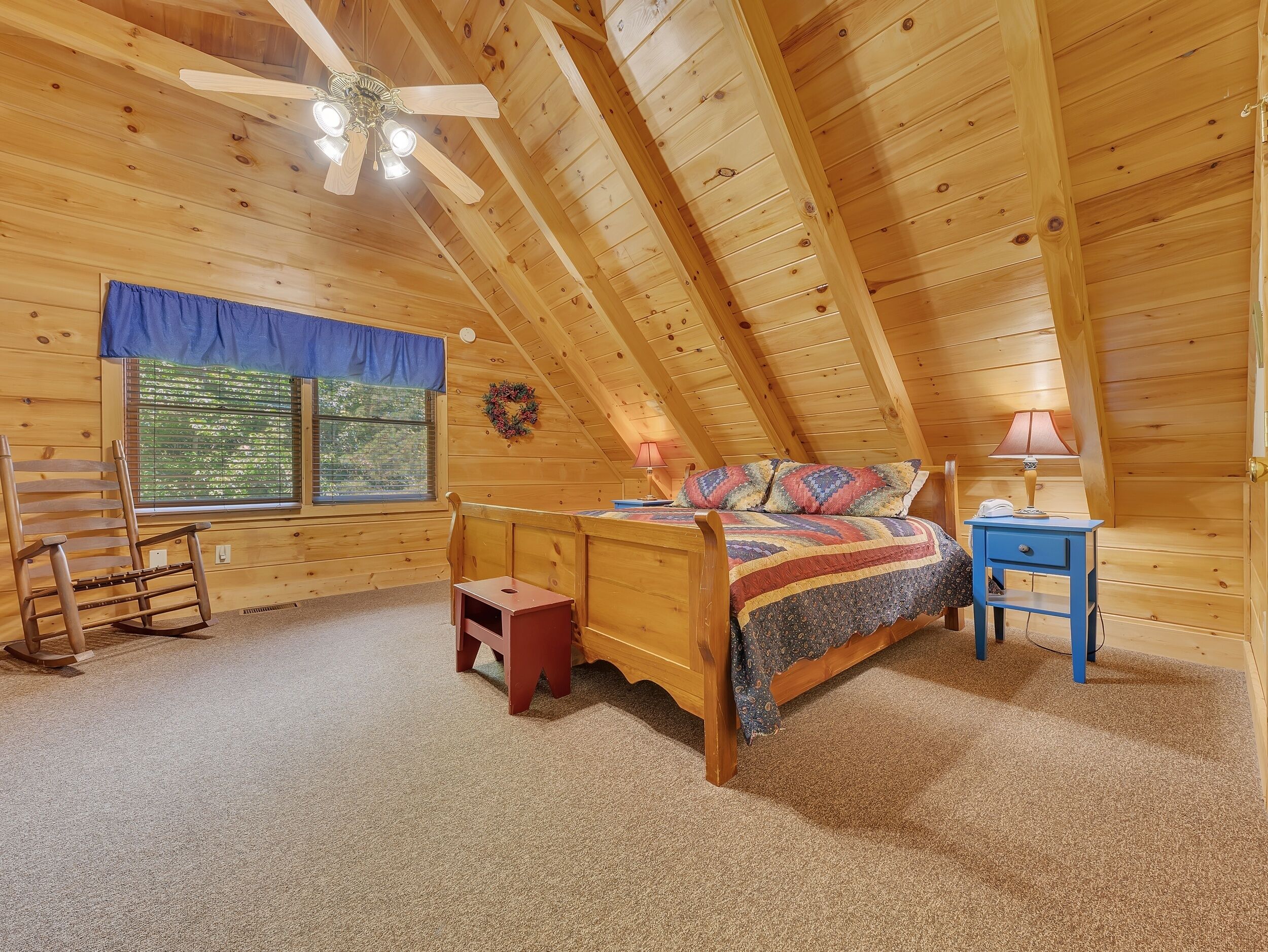 Une chambre confortable située sous les toits, avec un plafond en bois, un lit recouvert d’une literie colorée, une petite table de chevet, une chaise et une fenêtre donnant sur un paysage verdoyant.