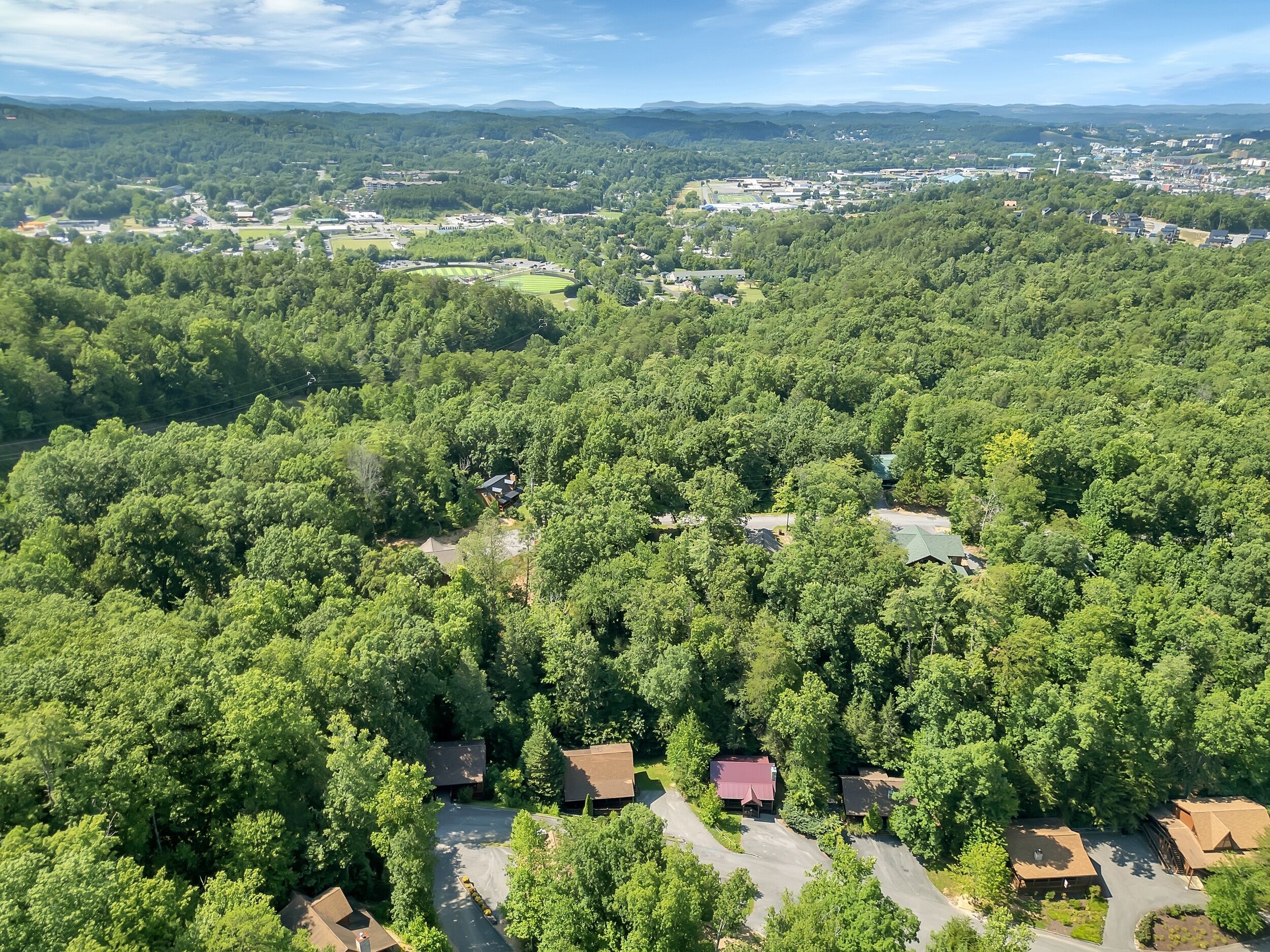 An aerial view of a residential area with multiple houses surrounded by dense forest, a clear sky, and distant buildings.