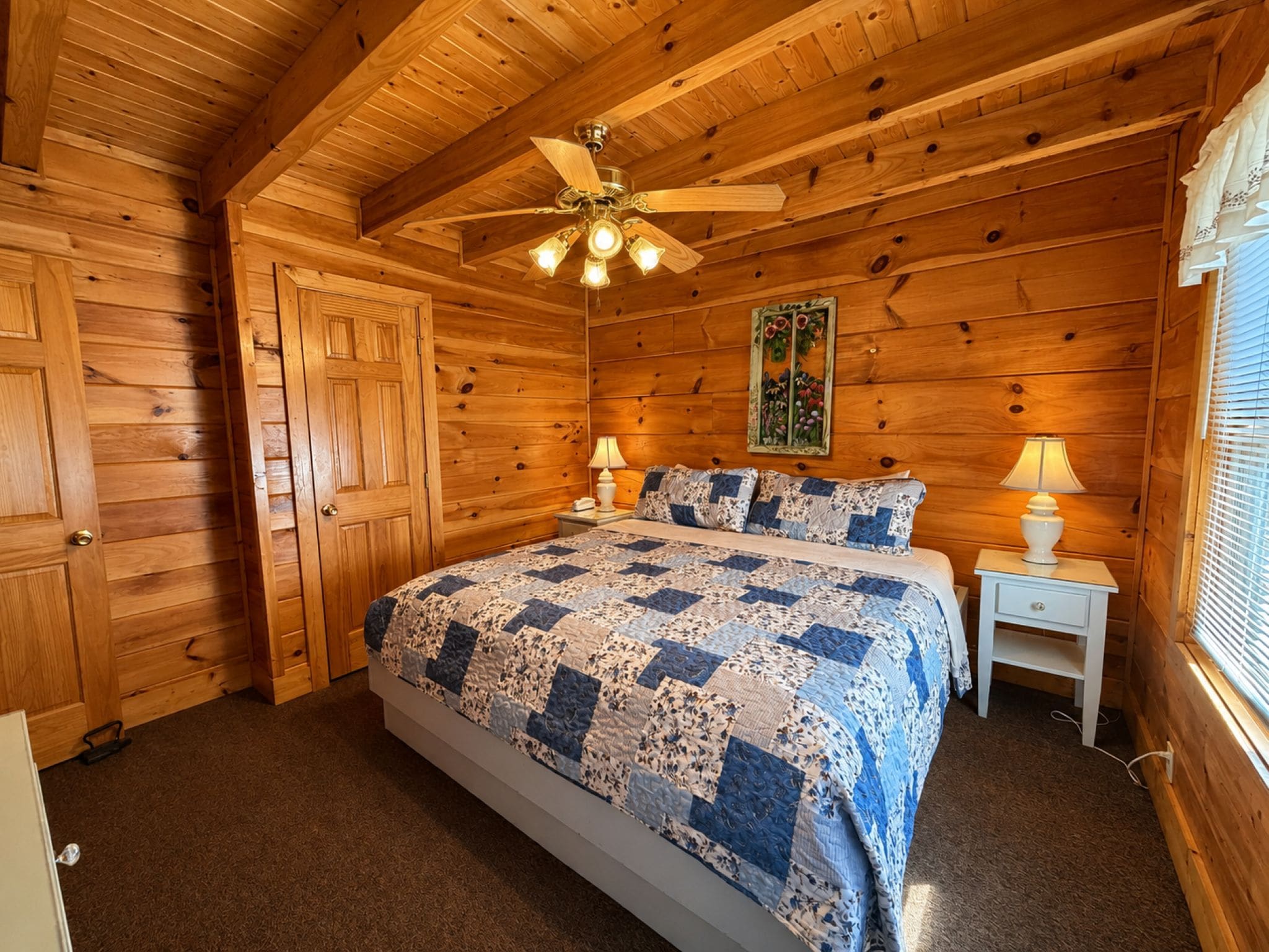 A wooden cabin bedroom with a bed, two bedside lamps, a ceiling fan, and a window with blinds.