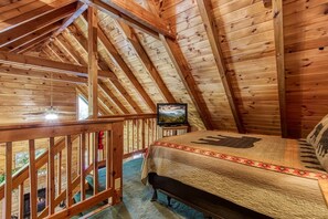 A cozy attic bedroom with a wooden ceiling, a bed with a patterned bedspread, a television on a wooden stand, and a view of a mountain landscape through a window. - Er5 - Log Heaven - Great Location! Close To All The Action! 1 Bedroom Cabin by RedAwning (Pigeon Forge)