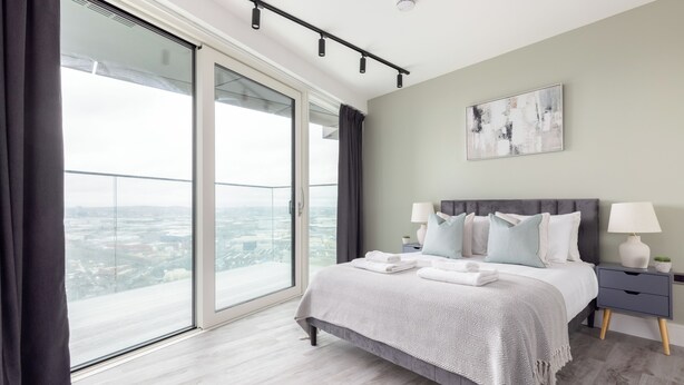 A modern bedroom with a large bed, a nightstand, a lamp, and a view of the city through a large window.