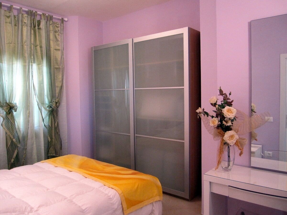 A bedroom with a bed, a wardrobe, a vanity with a mirror, and a vase with flowers.