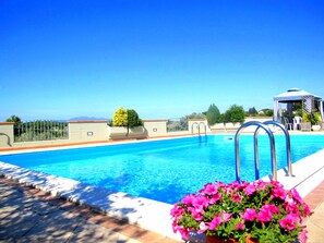 Une piscine à l’eau d’un bleu limpide, entourée d’une terrasse carrelée et d’une clôture en métal. On aperçoit des pots de fleurs avec des fleurs roses et un kiosque au fond.