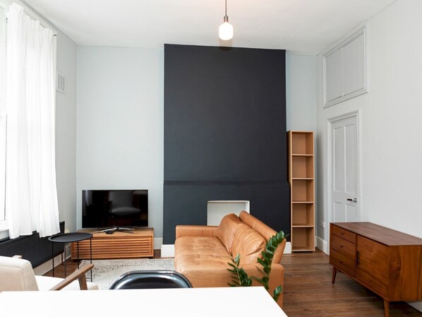 A modern living room with a large dark accent wall, a leather sofa, a wooden coffee table, a TV, and a bookshelf. - Charming and Spacious Apartment in Central Brixton - Pass the Keys (London)