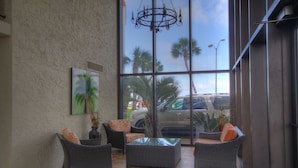 A sunlit indoor area with wicker furniture, a glass table, and a large window overlooking parked cars and palm trees.