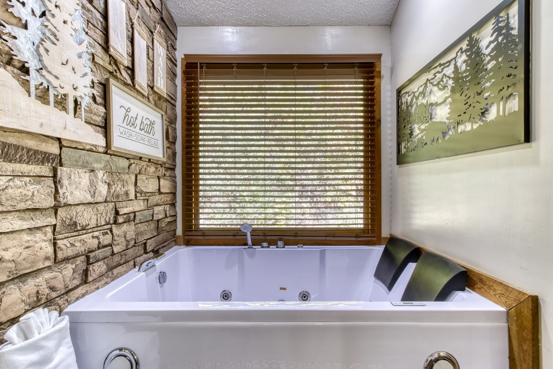 A bathroom with a stone wall, a window with blinds, a bathtub, and a framed picture on the wall.