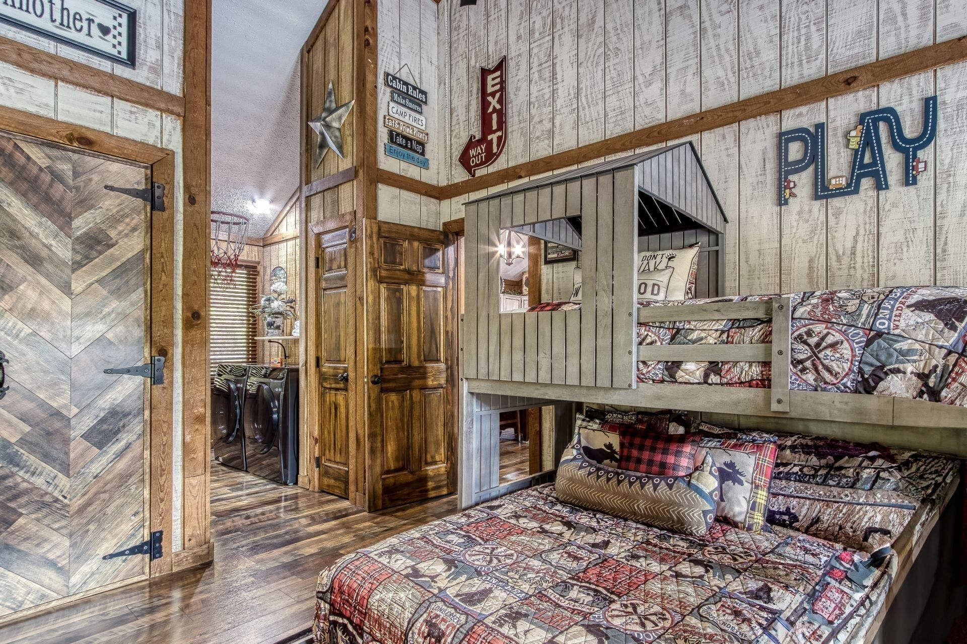A rustic cabin-style room with a herringbone floor, a bunk bed with patterned bedding, a wooden door, and cabin rules signs.