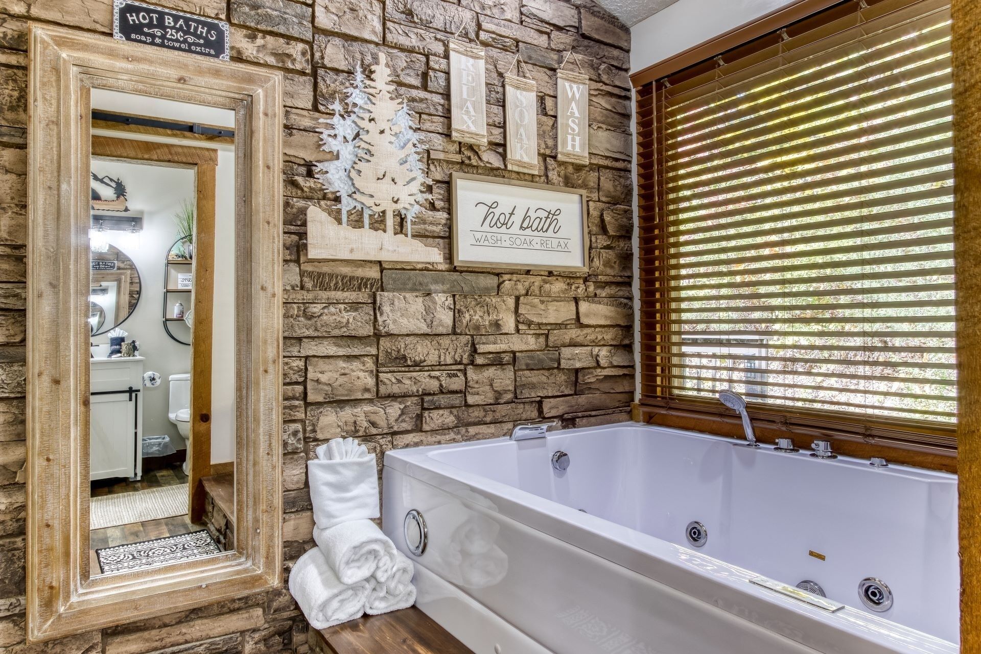 A bathroom with a stone wall, a framed sign reading "Hot Bath," a large tub, and a window with blinds.