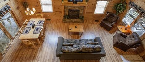 A cozy log cabin interior with a fireplace, a television mounted above it, a dining area with a table and chairs, and a seating area with a sofa and armchairs.