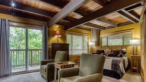A cozy room with a bed, two chairs, a small table, and a lamp. There is a large window with blinds and a view of trees outside. - Gatlinburg Views A Unique Cabin Mountain Views Expansive Decks Adjacent To Gatlinburg & Ski Lodge 1 Bedroom Cabin by RedAwning (Gatlinburg)