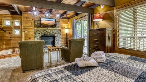 A cozy log cabin room with a fireplace, two armchairs, a small table, and a bed with a checkered blanket. - Gatlinburg Views A Unique Cabin Mountain Views Expansive Decks Adjacent To Gatlinburg & Ski Lodge 1 Bedroom Cabin by RedAwning (Gatlinburg)