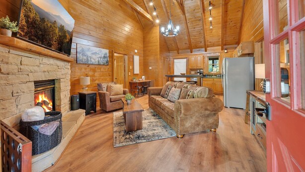 Un intérieur de chalet chaleureux avec une cheminée en pierre, un plancher en bois, un espace de vie avec des canapés et une table basse, une cuisine avec des armoires en bois et un téléviseur fixé au mur.