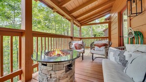 Une terrasse en bois couverte, dotée d’un foyer en pierre, de meubles en osier et offrant une vue sur la forêt environnante.