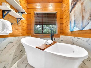 Une salle de bain moderne avec une baignoire sur pieds, des étagères en bois et une fenêtre avec des stores.