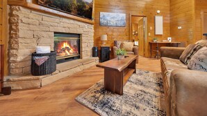 L’intérieur chaleureux d’un chalet en bois, avec une cheminée en pierre, des murs en bois et une grande fenêtre donnant sur un paysage boisé.